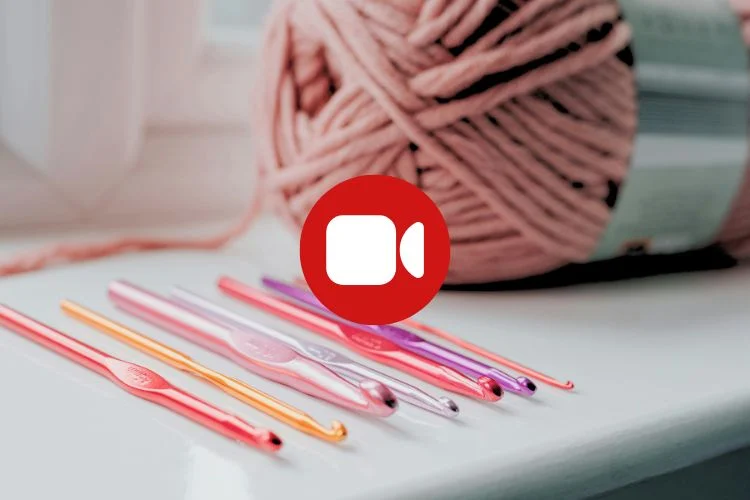 A row of colorful crochet hooks lies on a white surface with a large skein of pink yarn in the background; a video camera icon is overlaid in the center.