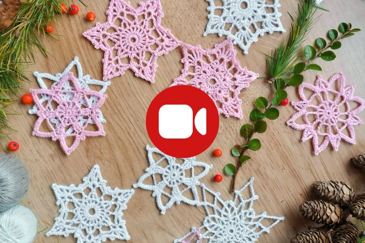 Crocheted snowflake ornaments in pink and white are arranged on a wooden surface with pine cones, greenery, red berries, and a yarn ball. A video call icon is overlaid in the center.