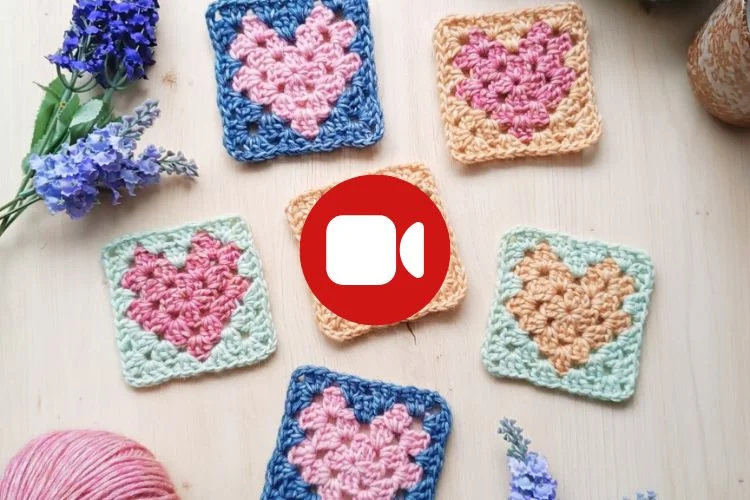 Six crocheted granny squares with heart designs are arranged on a wooden surface with flowers and yarn, and a red video call icon is centered over the image.