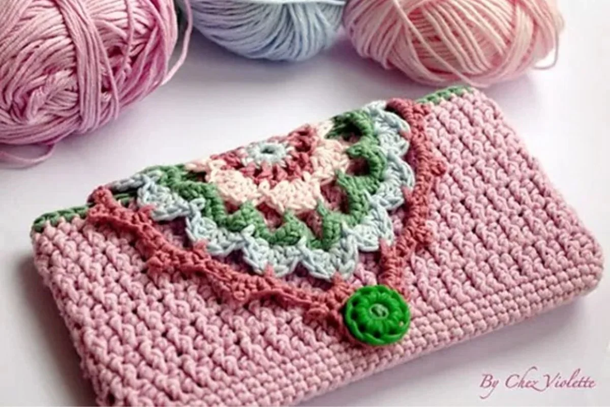 A crocheted pink pouch with a decorative flap in green, white, and pink yarn, finished with a green button. A charming crocheted snowman and two balls of yarn sit in the background.