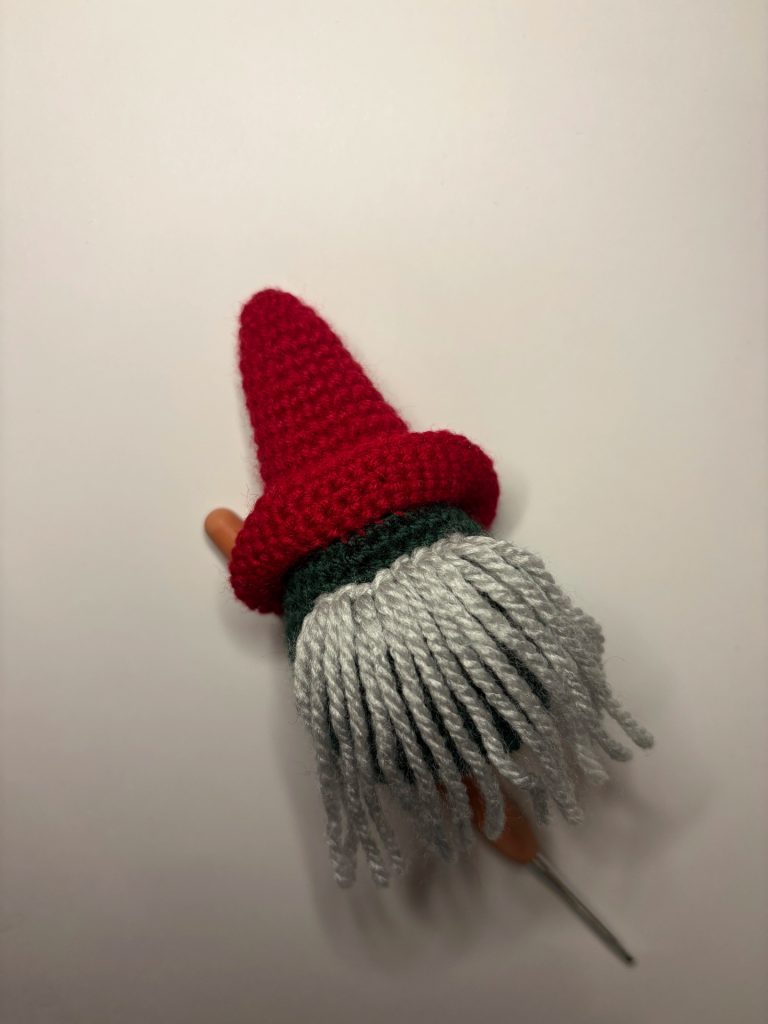 A crocheted gnome figure with a red hat, green body, and long gray yarn beard lies on a white surface—perfect for those who love tiny gnomes or want inspiration for their next pumpkin crochet pattern.