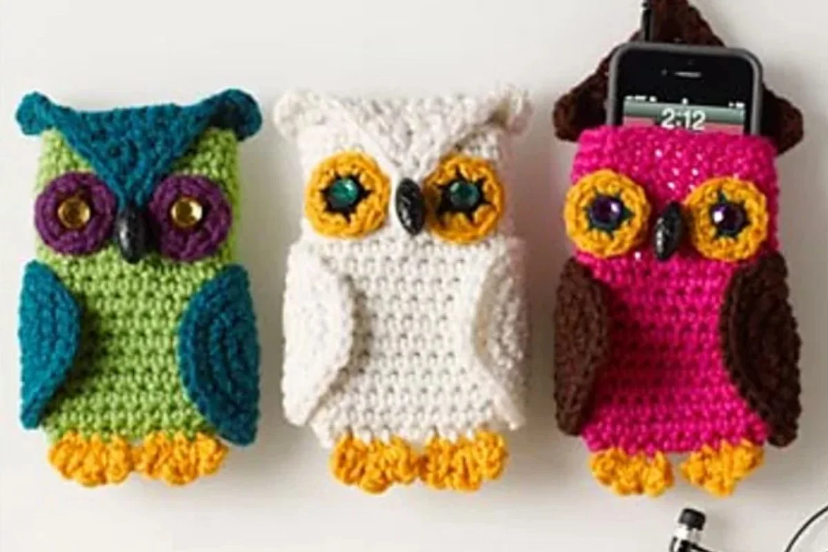 Three crocheted owl-shaped phone cases in green, white, and pink colors are displayed side by side, with a phone partially inserted into the pink case—a charming match for any crocheted snowman accessory.