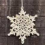 A cream-colored crocheted snowflake ornament, inspired by Winterlore, hangs by a string against a textured wooden background.
