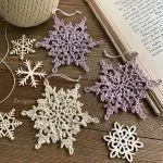 Crocheted and wooden snowflake ornaments are arranged on a wooden surface next to a ball of twine and an open book, capturing the spirit of Winterlore in cozy detail.