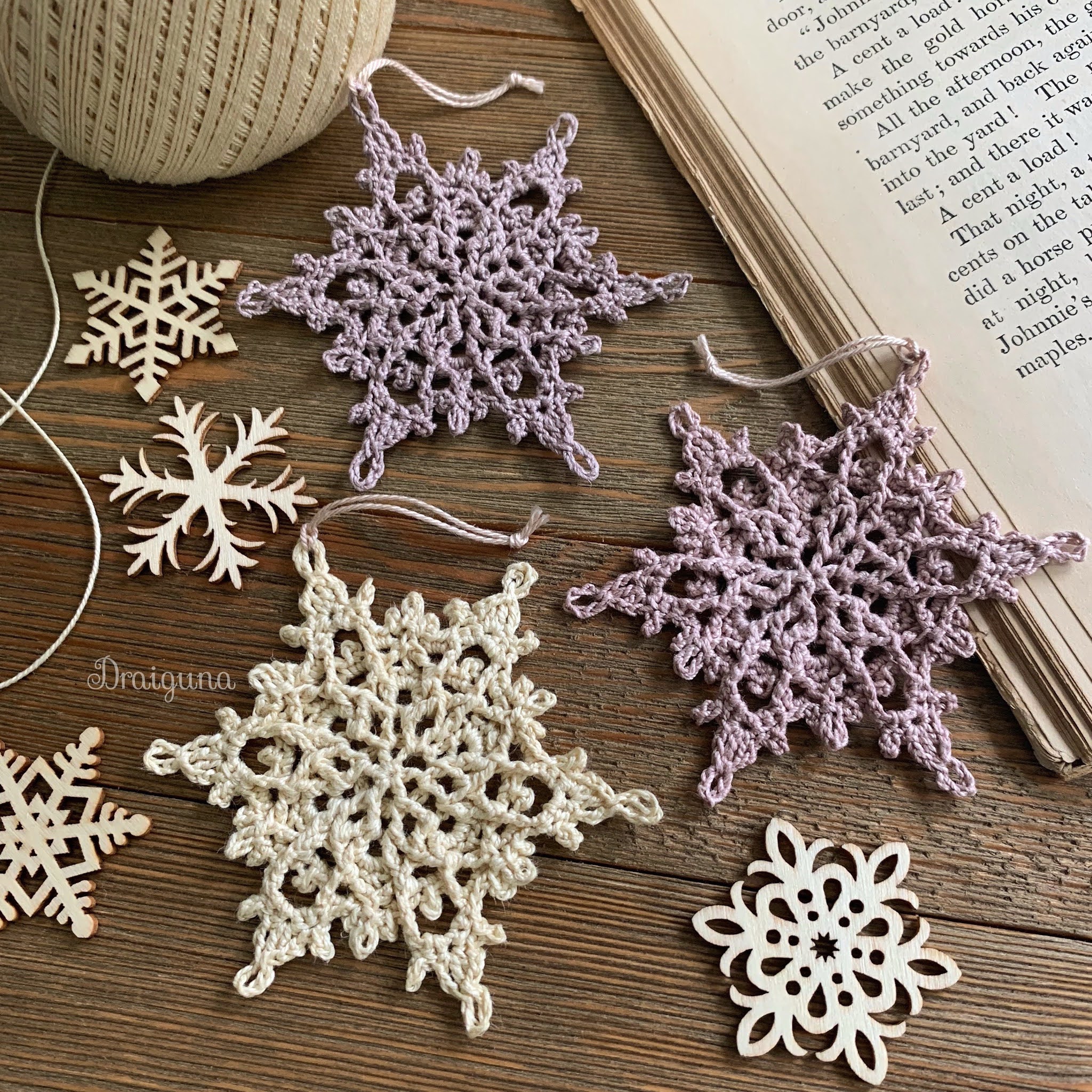 Crocheted and wooden snowflake ornaments are arranged on a wooden surface next to a ball of twine and an open book, capturing the spirit of Winterlore in cozy detail.