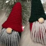 Two tiny crochet gnome ornaments with knit hats, gray and white yarn beards, and round noses are placed on a white surface with pine branches and string lights—perfect alongside your favorite crochet Christmas ornaments pattern.