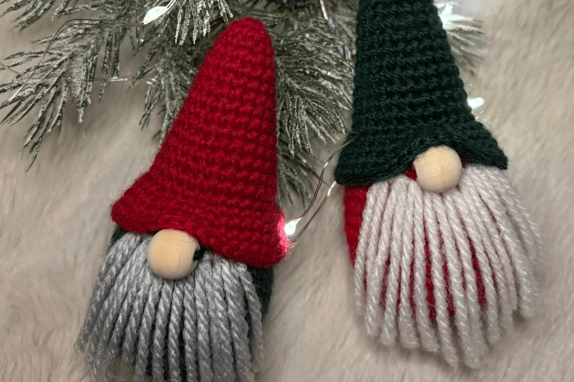 Two tiny crochet gnome ornaments with knit hats, gray and white yarn beards, and round noses are placed on a white surface with pine branches and string lights—perfect alongside your favorite crochet Christmas ornaments pattern.
