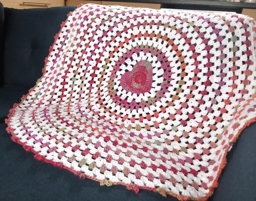 A multicolored crocheted wave baby blanket with a heart pattern in the center is draped over a dark blue sofa.