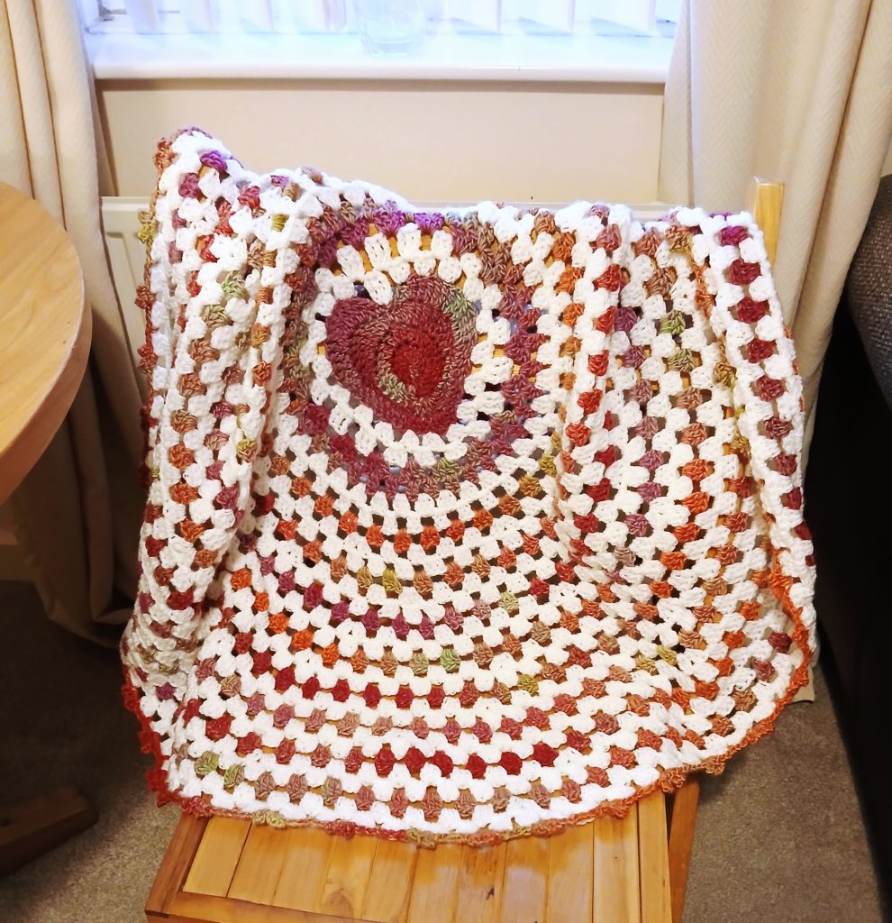 A wave baby blanket crocheted in a circular granny square pattern with red, orange, purple, and white yarn draped over a wooden chair.