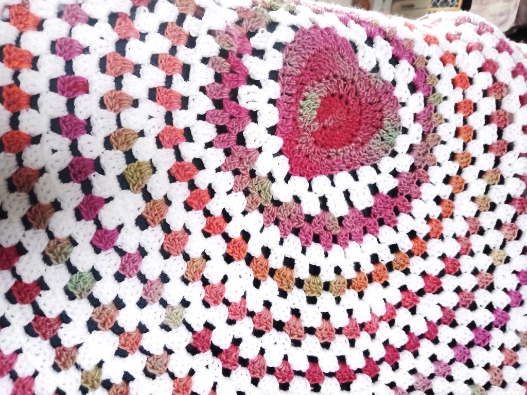 A crocheted wave baby blanket in white, pink, and orange granny squares features a large pink and green heart shape in one corner.
