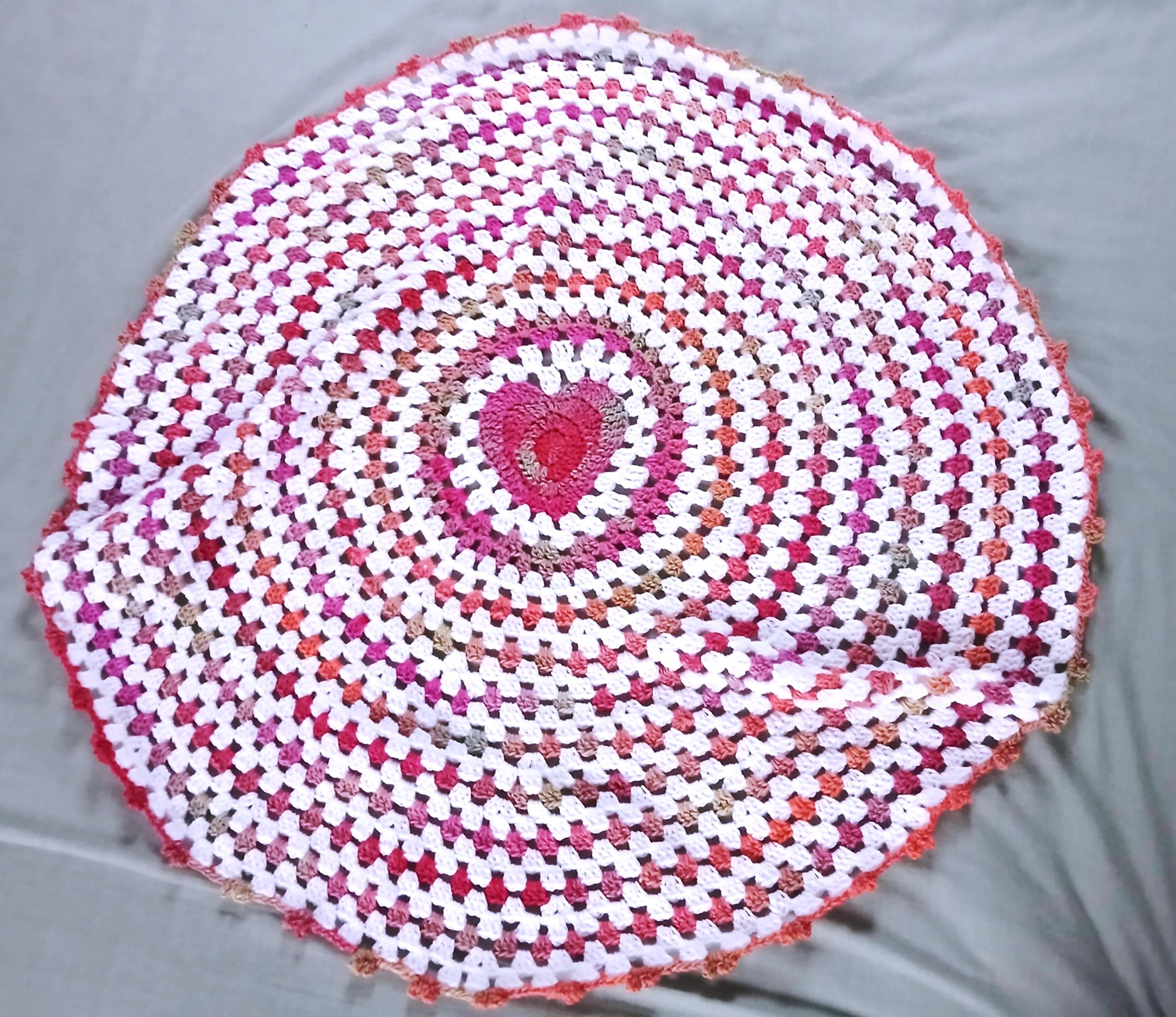 A round crocheted mat with a heart pattern in the center, inspired by wave baby blanket designs, features alternating rows of pink, white, red, and beige yarn on a light gray surface.