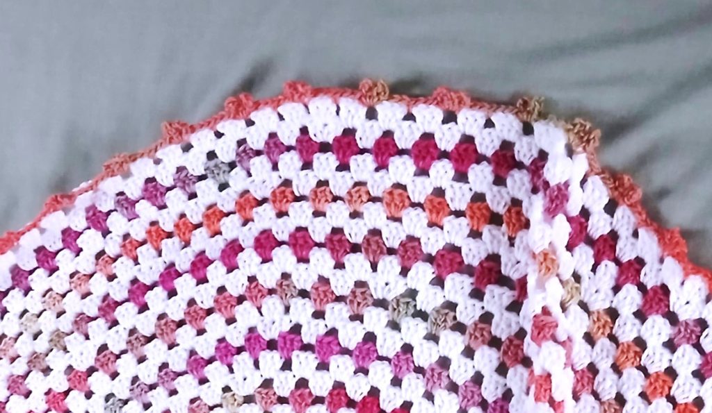 A close-up of a handmade crochet granny square wave baby blanket with rows of white, pink, red, orange, and brown yarn on a light grey background.