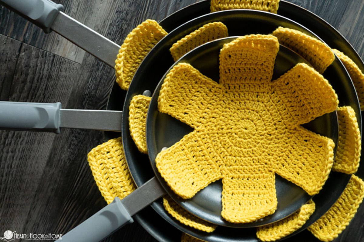 Three nonstick frying pans are stacked with yellow crocheted pan protectors placed between them on a dark wooden surface.
