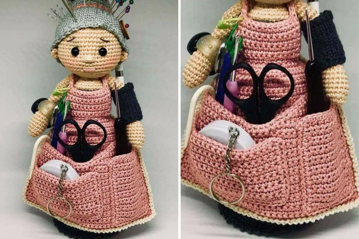 A crocheted doll wearing a pink apron holds sewing tools such as scissors, pens, thread, and pins in its apron pockets and on its head.