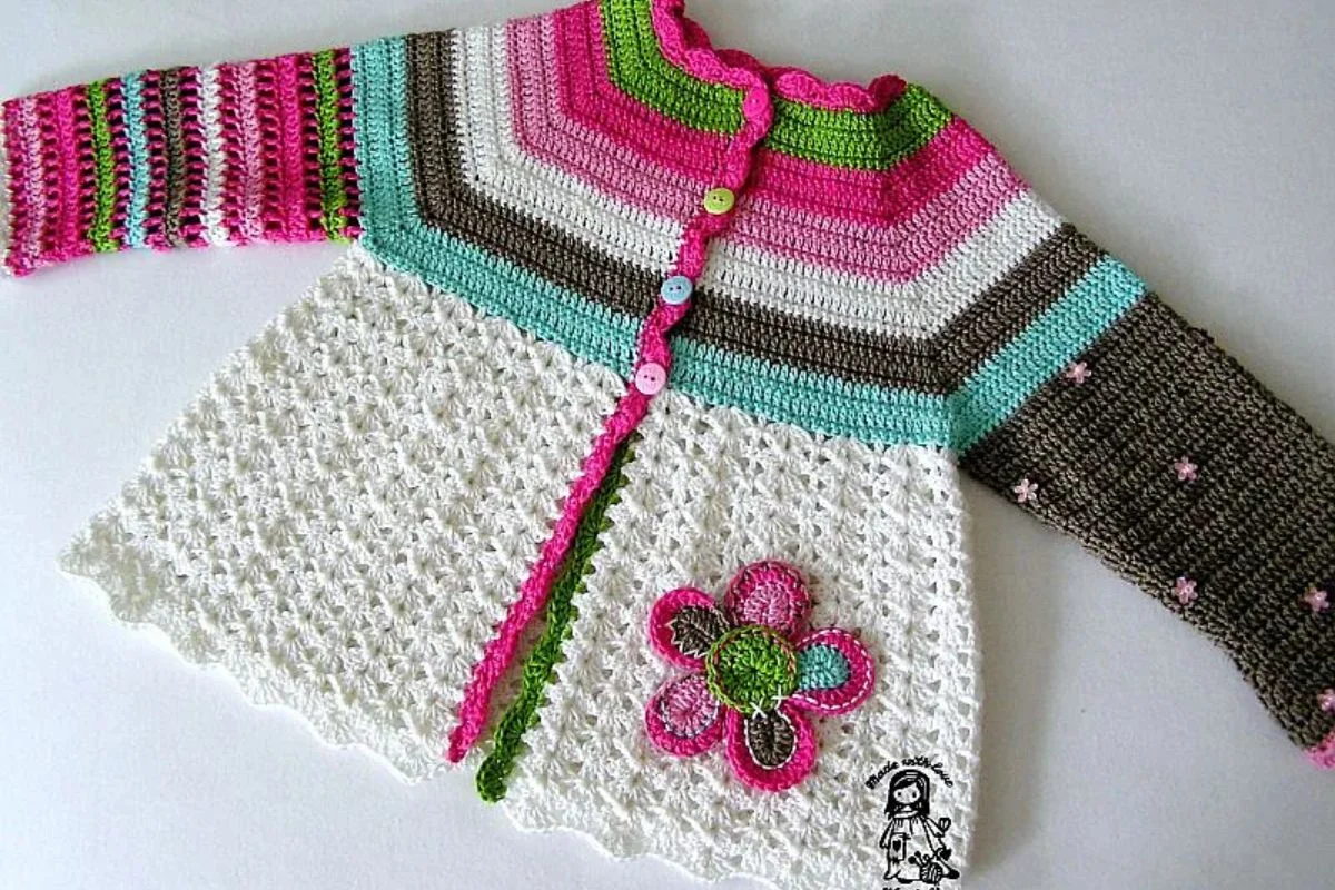 A colorful crocheted baby cardigan with pink, green, white, turquoise, and brown stripes, flower applique, and multicolored buttons, laid flat on a white surface.