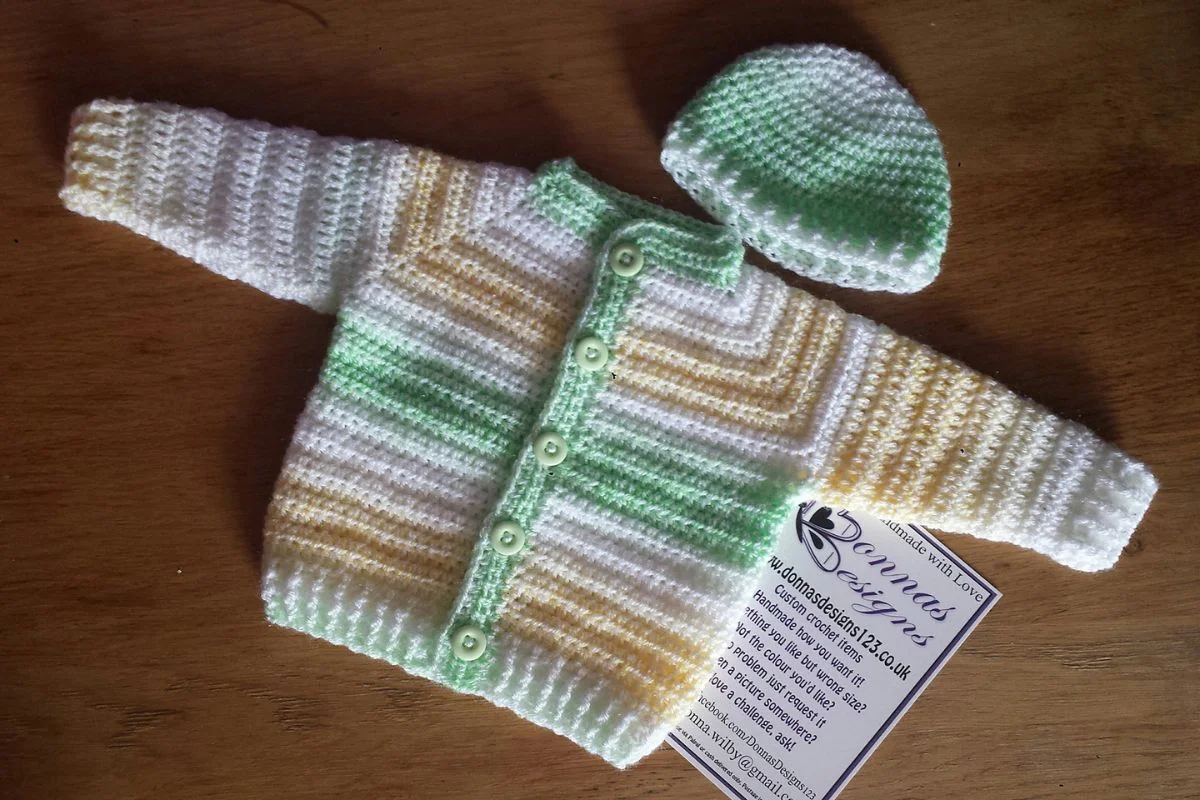 A Top Down Baby Jacket and matching hat in white, green, and yellow stripes are displayed on a wooden surface with a business card partially visible underneath.
