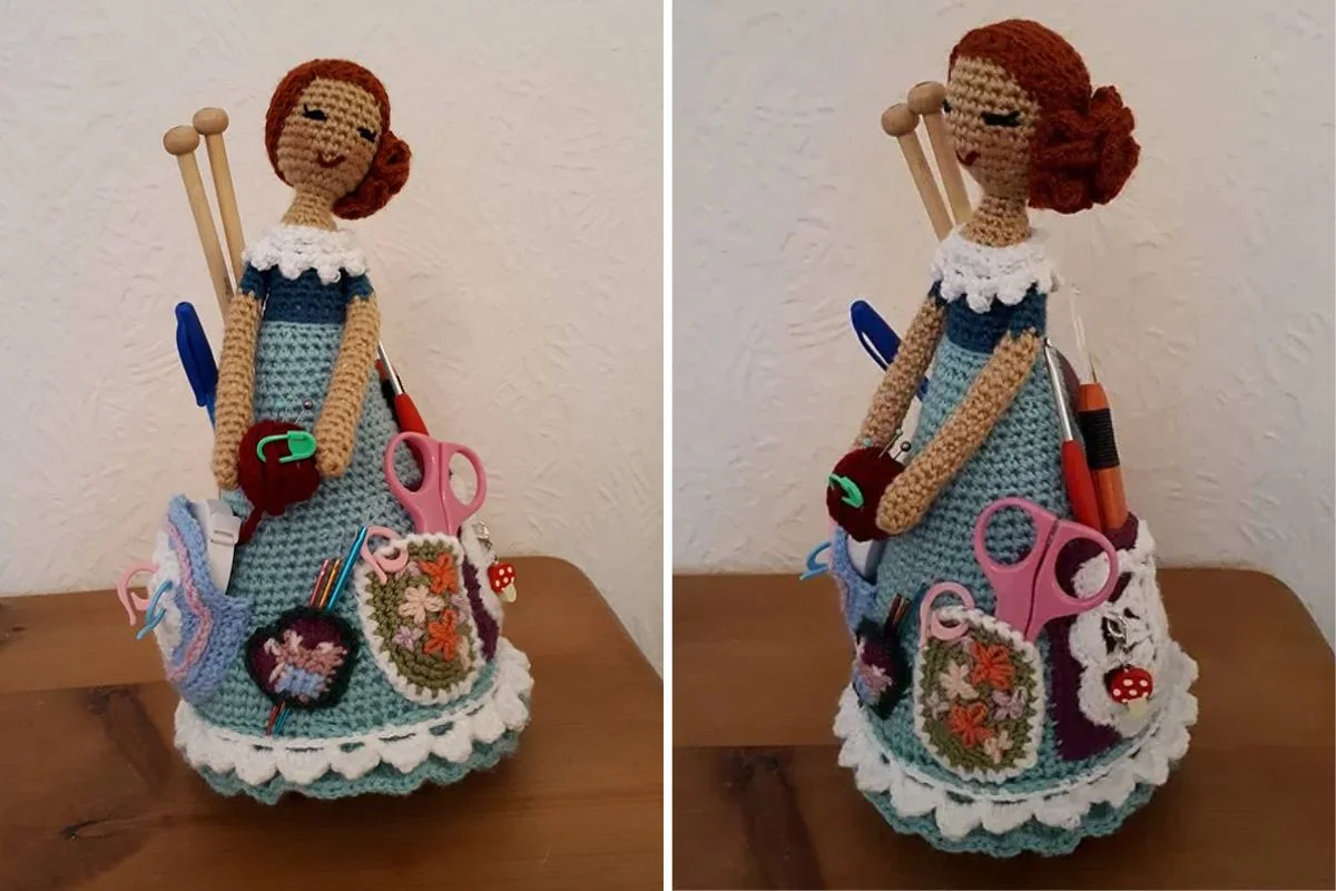 A crocheted Crafter Granny doll holds knitting needles, pens, and scissors in pockets on its skirt, serving as a decorative organizer on a wooden surface.