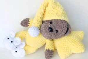 A plush crochet teddy bear in pajamas and a yellow nightcap lies on its side, cuddling a yellow star pillow and wearing white mouse-shaped slippers.