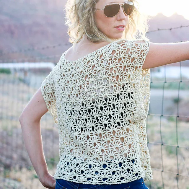 A woman in sunglasses and a lacy, cream-colored crochet boho top stands outdoors near a wire fence, her handmade wardrobe on display as she faces away from the camera.