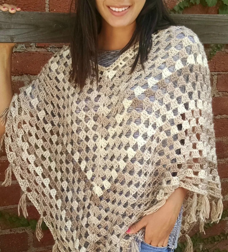 Woman wearing a crocheted poncho with neutral colors and fringe, standing against a brick wall and holding a wooden beam—perfect inspiration for your Handmade Wardrobe or those seeking stylish Crochet Boho Tops.