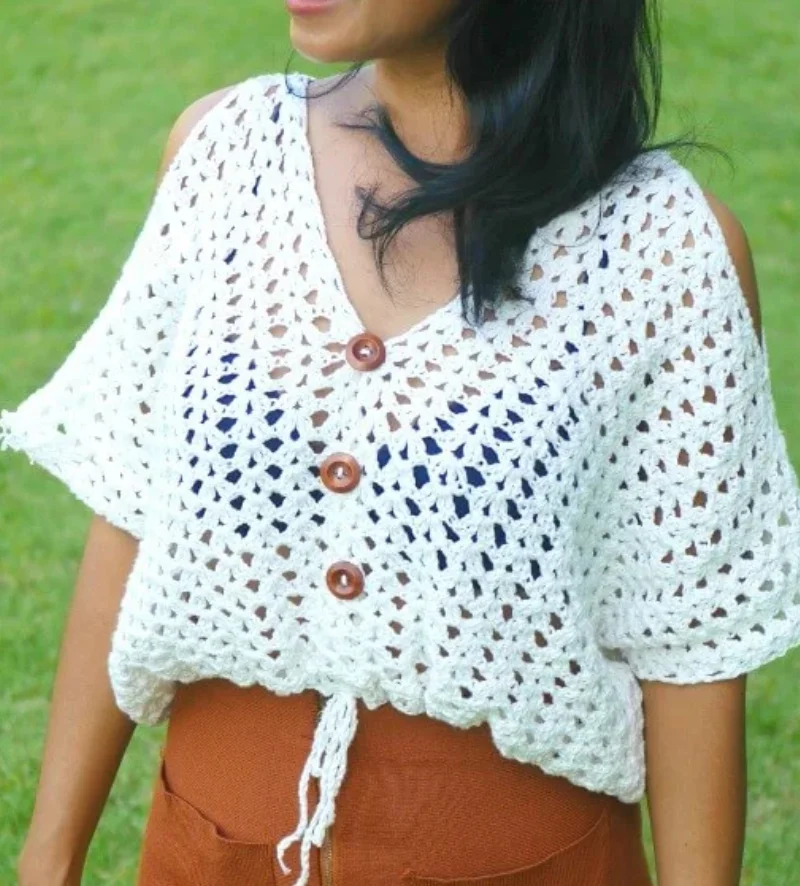 A person wearing a white crochet boho top with brown buttons and a drawstring waist, paired with rust-colored pants, stands outdoors on grass—perfect inspiration for your handmade wardrobe.