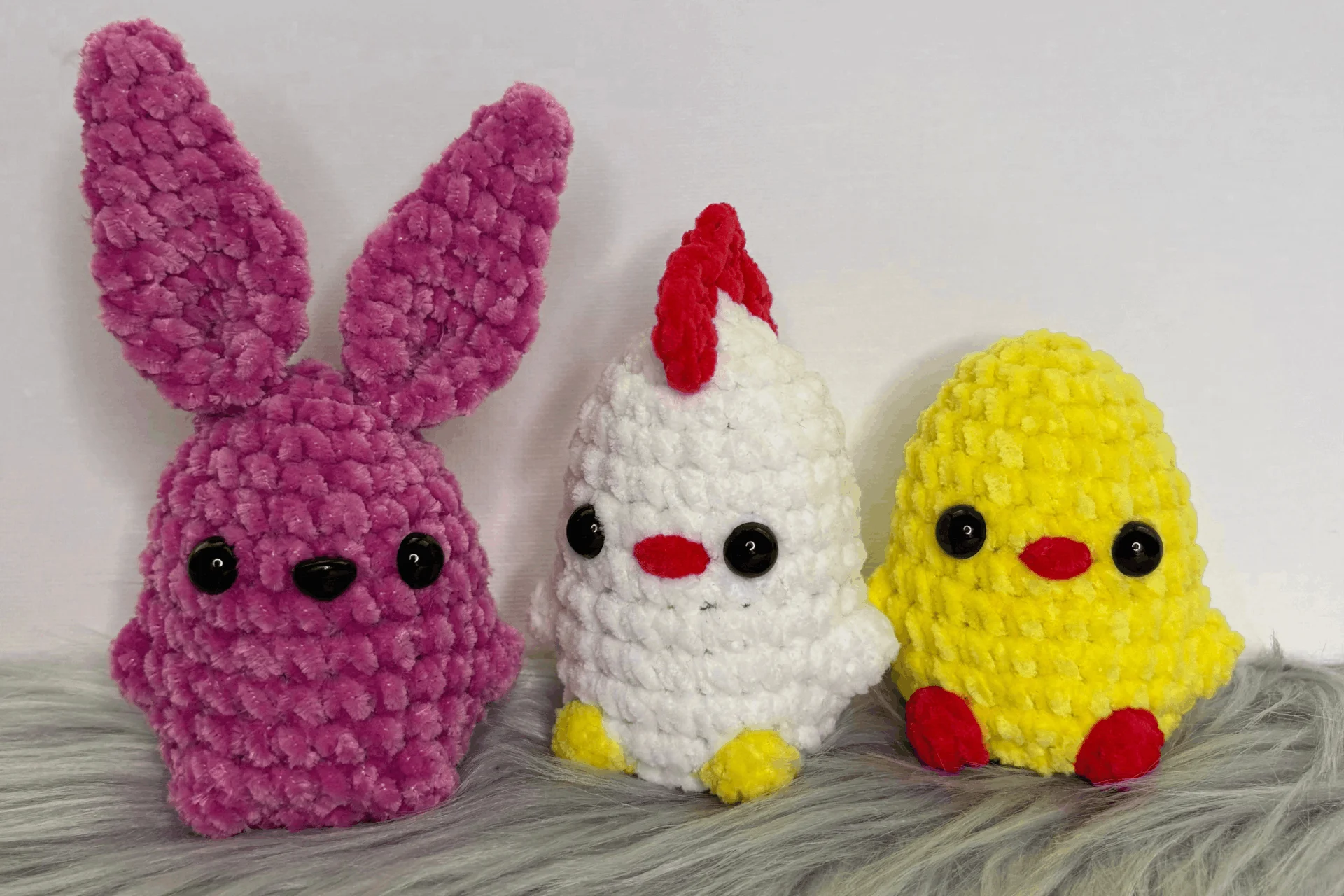 Three crocheted plush toys—a purple bunny, a white chicken, and a yellow chick—are displayed on a faux fur surface against a plain background, perfect companions for your next free crochet bunny basket pattern project.