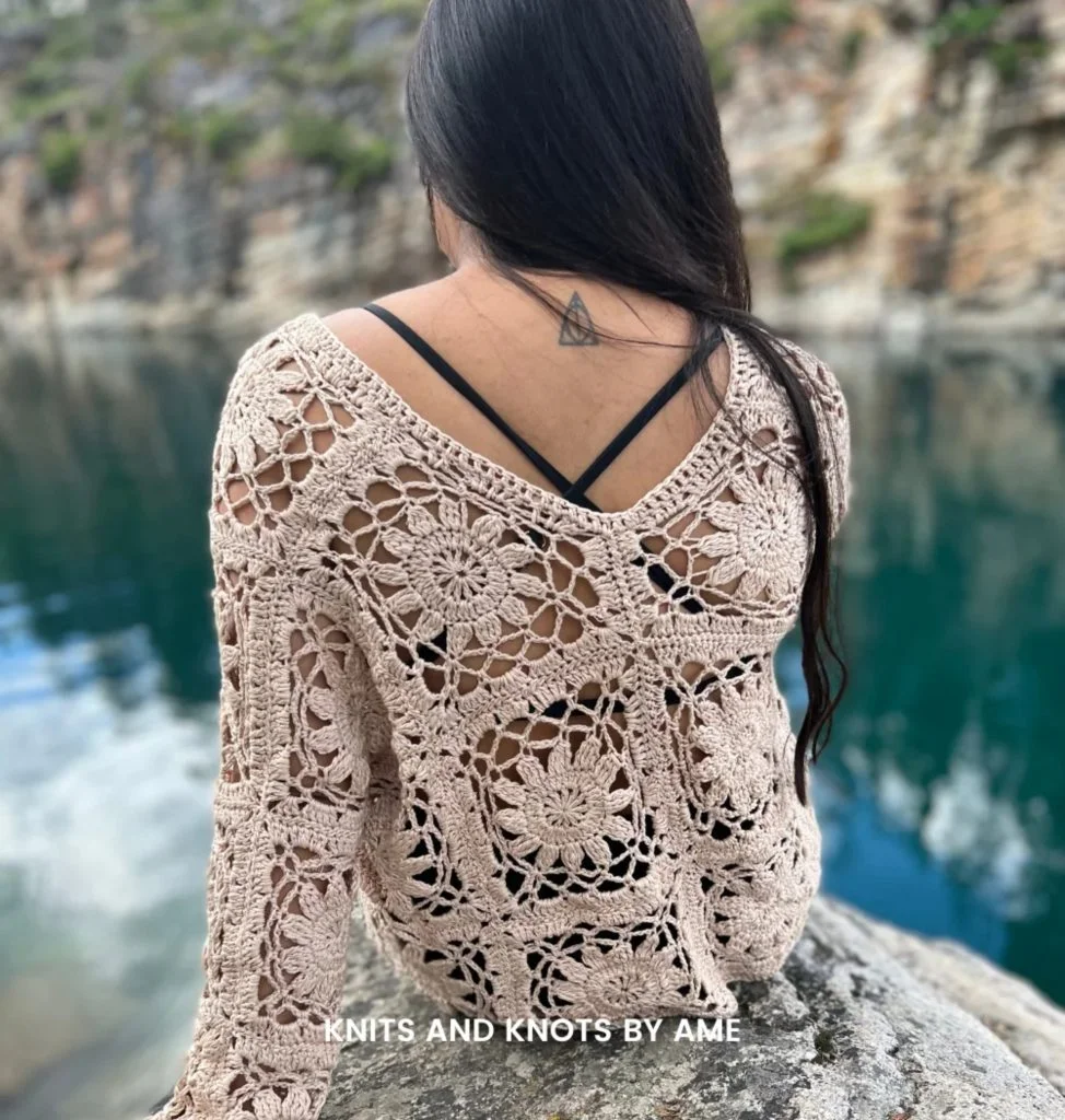 A person with long dark hair sits on a rock by a body of water, wearing a beige crochet boho sweater over a black top. Text reads "KNITS AND KNOTS BY AME.