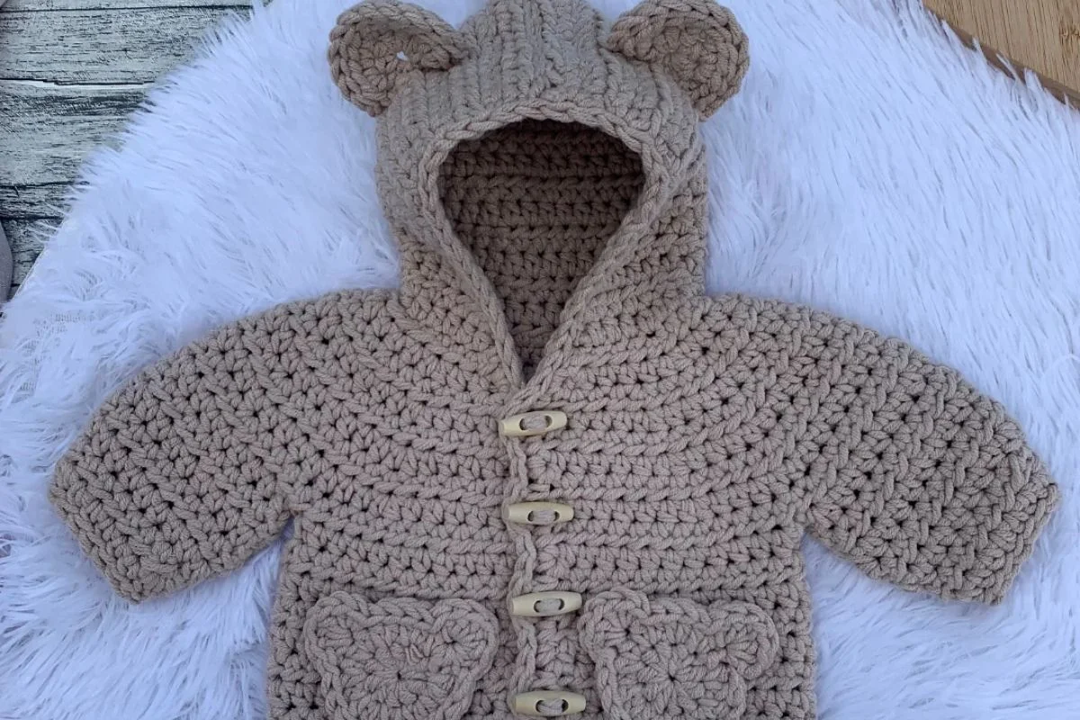A beige crocheted baby sweater with a hood featuring bear ears, wooden buttons, and two front pockets, displayed on a white textured surface.