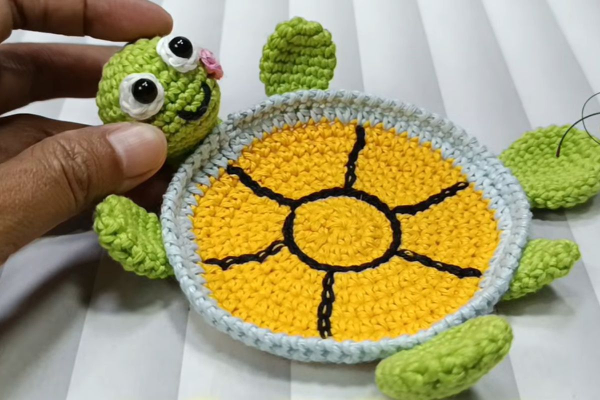 A hand holds a crocheted turtle head next to a yellow and green crochet turtle coaster body laying on a white surface.