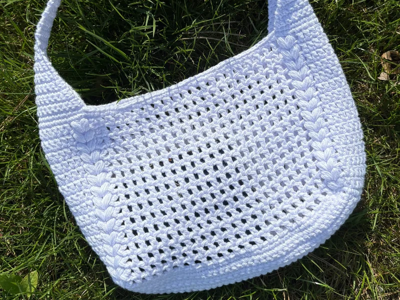 A white summer bag with a textured crochet pattern lies on green grass, perfect for those who love a stylish touch—pair it with your favorite outfit for a seasonal accent.