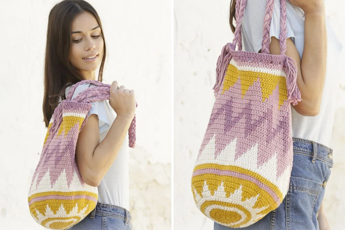 A woman wearing a white T-shirt and denim shorts carries an Andes Sunrise Bag, a large, colorful crocheted shoulder bag with pink, yellow, and white geometric patterns.