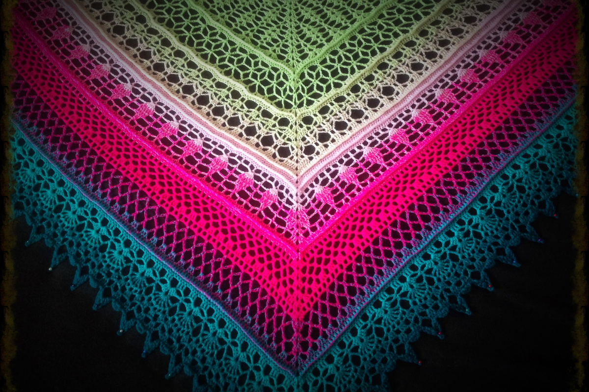 A multicolored, triangular Edlothia Shawl with intricate lace patterns in shades of green, yellow, pink, purple, and blue is displayed against a dark background.
