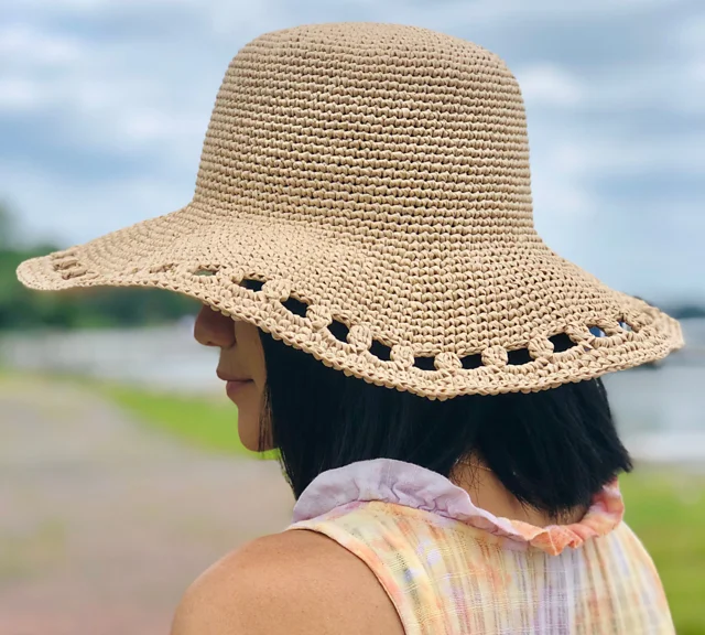 A person with shoulder-length black hair wears a wide-brimmed, woven straw hat and a sleeveless top, standing outdoors near water on a cloudy day.