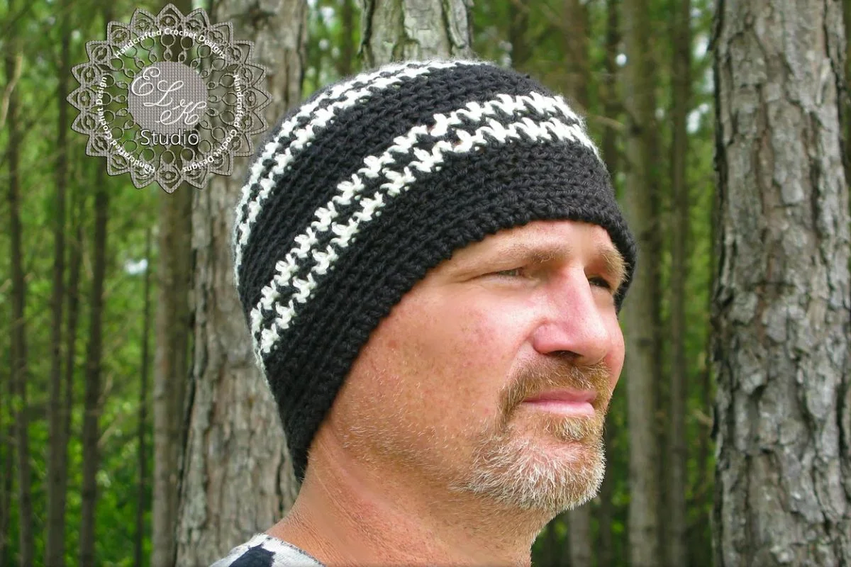 A man with a short beard wears a black and white striped crocheted beanie, standing outdoors in front of tall trees.