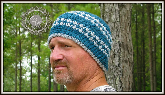 A man with a short beard wears a blue and gray striped crochet beanie, standing outdoors among tall trees as dusk till dawn light filters through the leaves.