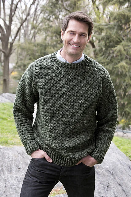 A man standing outdoors wearing a dark green knit sweater and black jeans, smiling with his hands in his pockets. Trees and rocks are visible in the background, capturing a peaceful Dusk Till Dawn vibe.