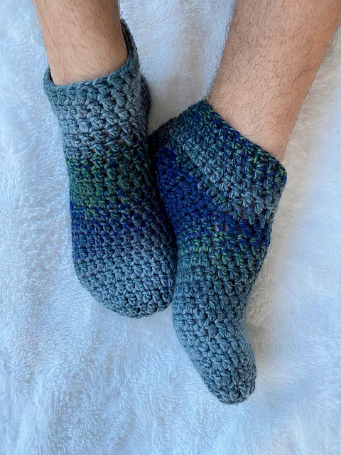Two feet wearing thick, crocheted slippers in blue and gray tones rest on a soft white surface, offering cozy comfort from dusk till dawn.