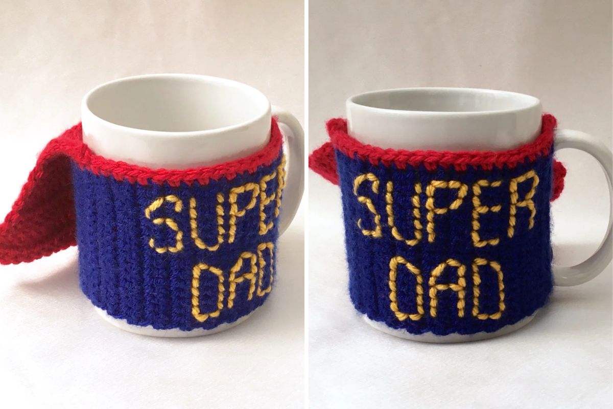 A white mug with a blue crochet cozy that says "SUPER DAD" in yellow letters and features a red cape attached to the handle.
