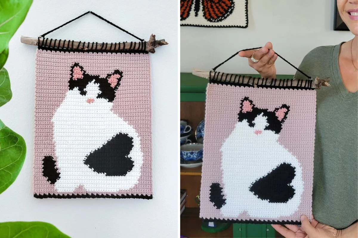 A crocheted Sweetheart Kitty Tapestry featuring a black and white cat on a light pink background, displayed on a stick and held up by a person.