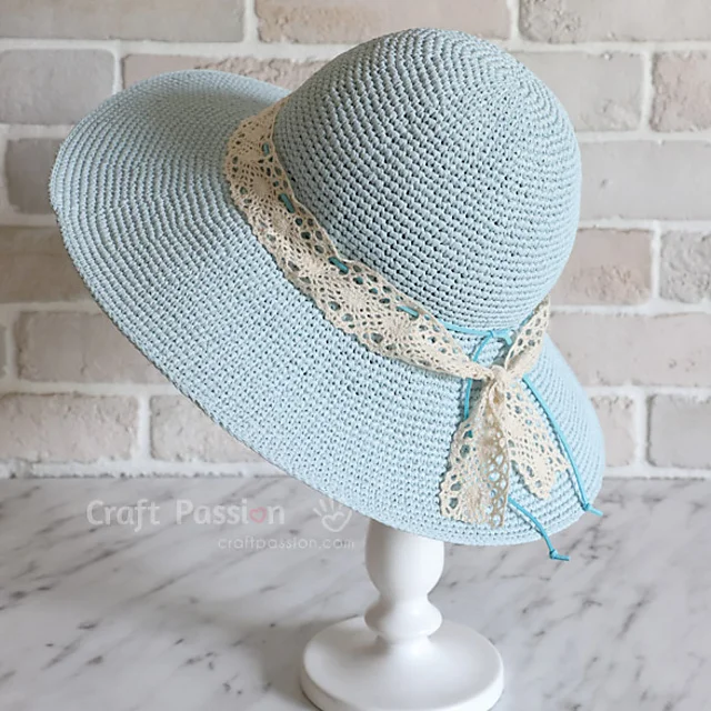 A light blue crocheted sun hat with a wide brim and a cream-colored lace ribbon, displayed on a white stand against a brick wall background.