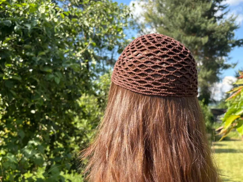 Person with long brown hair wearing a brown crochet mesh cap, standing outdoors with trees and greenery in the background.