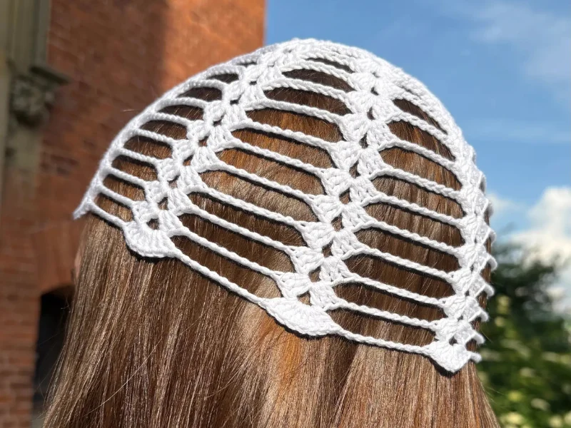 A person with long brown hair is wearing a white crochet mesh cap outdoors, with a brick building and sky in the background.