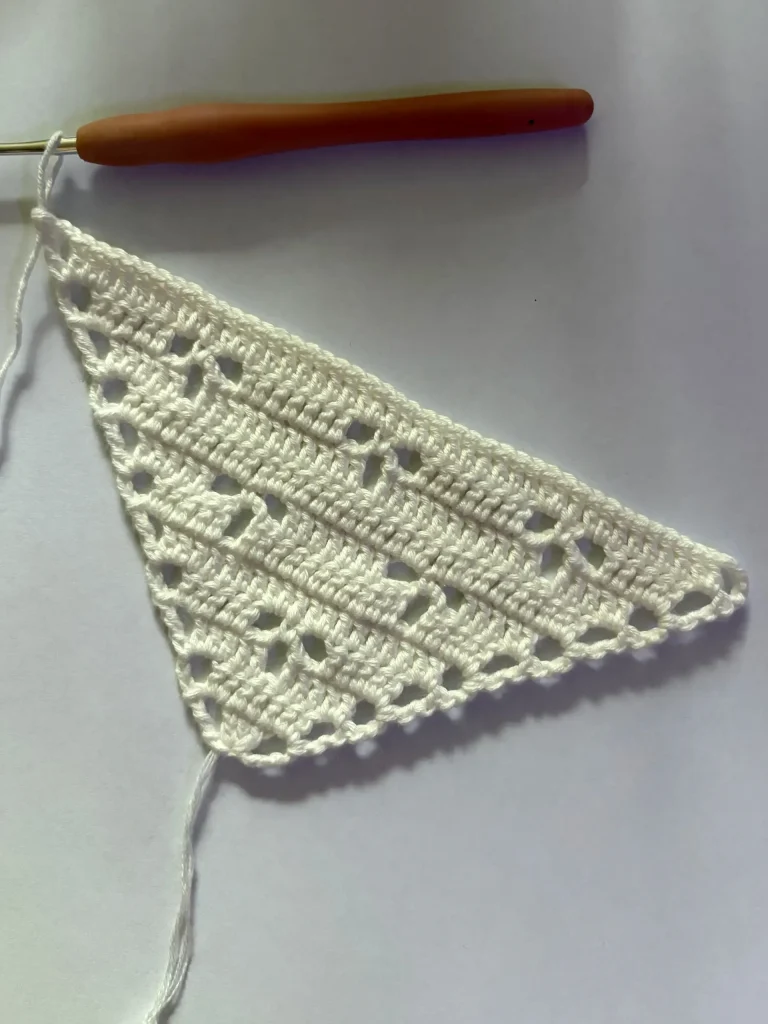 A white crocheted triangular piece with small gaps, resembling a delicate crochet bandana, is attached to a wooden-handled crochet hook on a plain background.