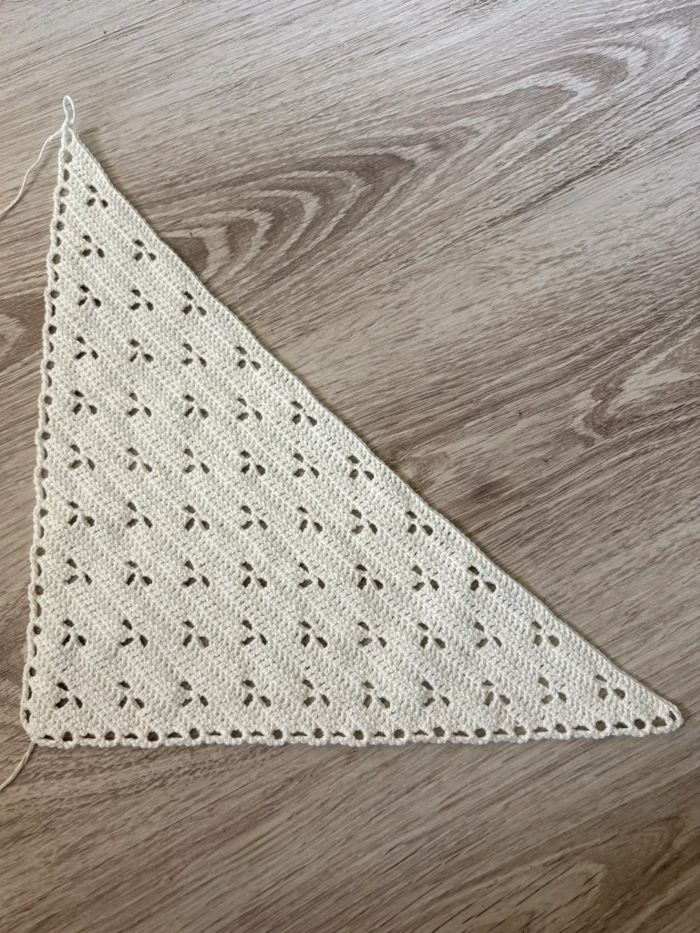 A triangular knitted or crocheted swatch with a pattern of small holes, reminiscent of a delicate crochet bandana, placed on a light wooden surface.