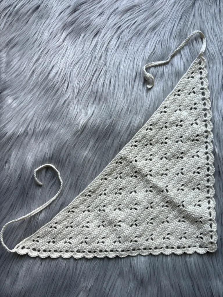 A cream-colored crocheted triangle scarf with a scalloped edge and ties, laid flat on a gray faux fur surface, evokes cozy autumn vibesโperfect for pairing with your favorite pumpkin crochet pattern accessories.
