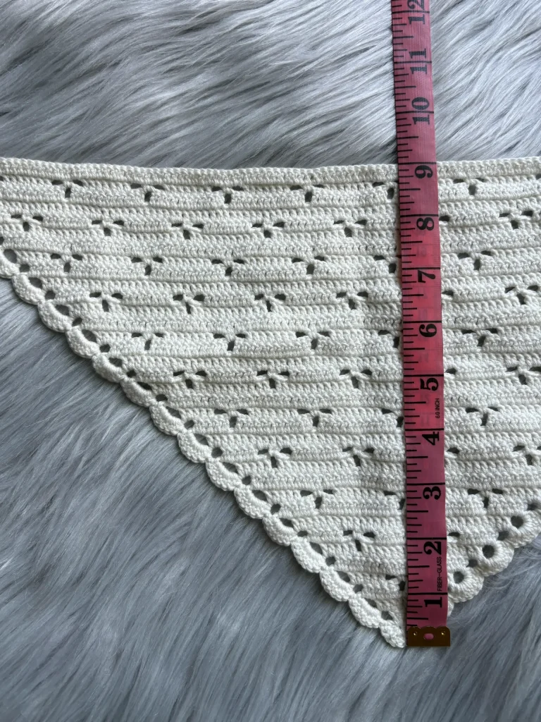 A pink tape measure is placed vertically next to a crocheted triangular crochet bandana with scalloped edges, laid on a gray faux fur surface.