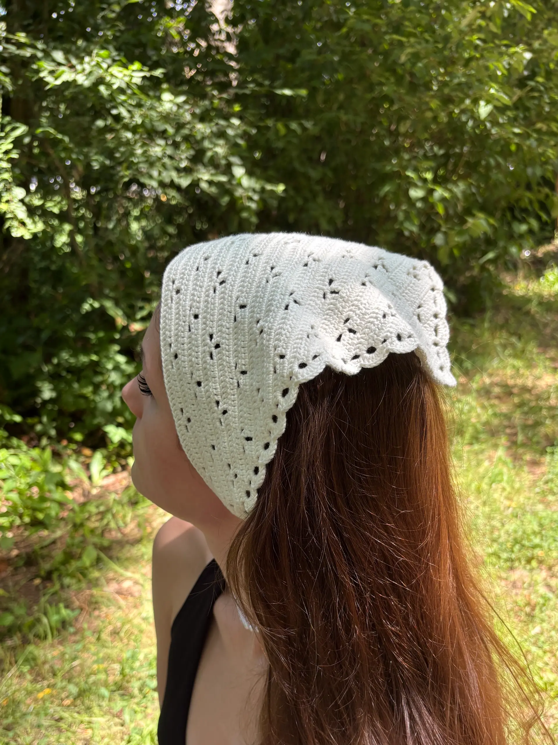A person with long brown hair wears a white crocheted headscarf, while standing outdoors in a green, leafy area.
