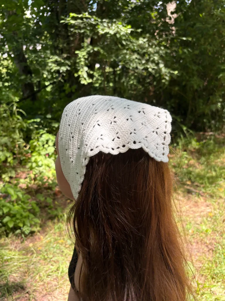 A person with long brown hair wears a white, crocheted headscarf, while standing outdoors in a sunlit, wooded area.