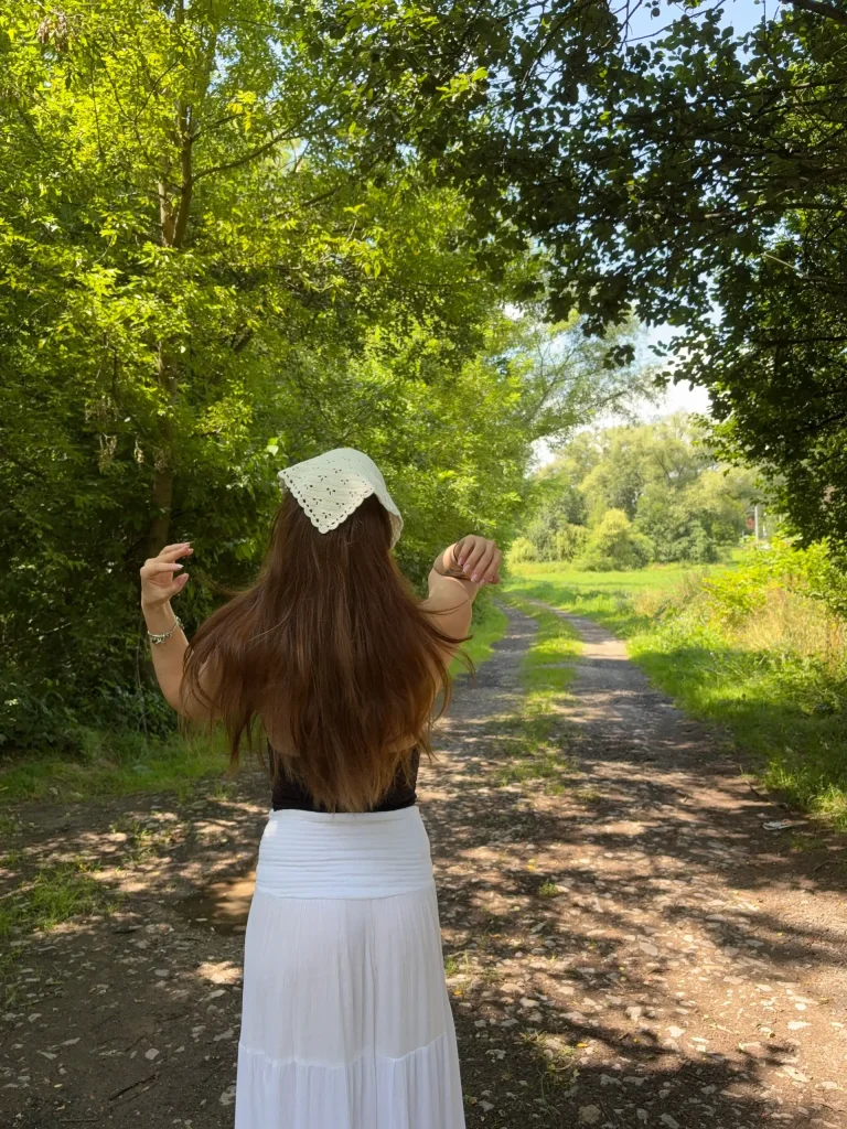 A person with long brown hair and a white headscarf stands on a sunlit dirt path in a green, wooded area, facing away from the camera. The peaceful scene captures an autumnal charmโperfect inspiration for anyone searching for a cute crochet summer pattern to try this season.