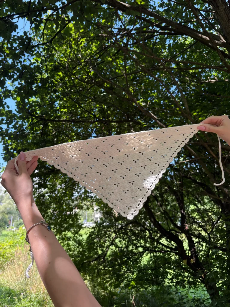A person holds up a crocheted white triangular bandana outdoors, with sunlight shining through the openwork pattern and trees in the backgroundโperfect inspiration for anyone searching for a summer crochet pattern.